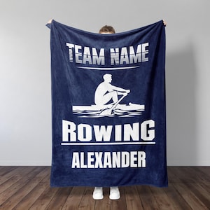 May include: A navy blue fleece blanket with a white silhouette of a rower in a boat. The text "TEAM NAME ROWING ALEXANDER" is printed in white on the blanket.