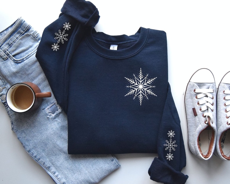 May include: A navy blue sweatshirt with a white snowflake design on the front and two smaller snowflakes on each sleeve. The sweatshirt is laid out on a white surface with a pair of gray canvas shoes and a pair of blue jeans.