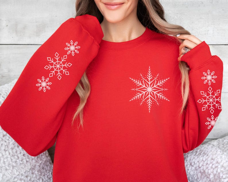 May include: Red sweatshirt with white snowflake designs on the sleeves and chest. The snowflakes are different sizes and have a simple, geometric design.