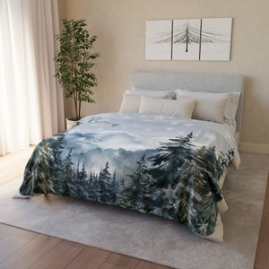 May include: A soft, blue and green fleece blanket with a watercolor design of a snowy forest. The blanket features a white background with a subtle snowflake pattern and the word "Cozy" written in cursive.