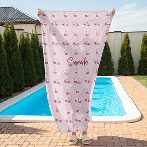 May include: A pink beach towel with a repeating pattern of red cherries. The towel has the name "Sarah" written in pink cursive lettering.