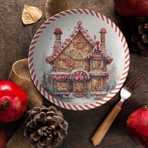 May include: A white ceramic plate with red and white candy cane stripes around the edge. The center of the plate features a watercolor illustration of a gingerbread house with candy details. The house is surrounded by green trees and snow.
