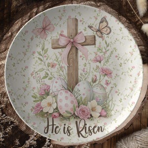 May include: A white ceramic plate with a floral design featuring a wooden cross with a pink ribbon, butterflies, and Easter eggs. The text "He is Risen" is printed on the plate.