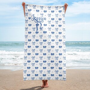 May include: White beach towel with a repeating pattern of blue bows and hearts. The name "Jessica" is written in cursive script in the center. The towel is held up on a beach with the ocean in the background.