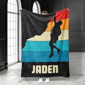 May include: A black fleece blanket with a retro-style design featuring a silhouette of a person rock climbing. The design includes a mountain, a rope, and a sunset. The blanket has the name "Jaden" printed in white letters at the bottom.