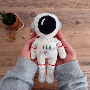 Könnte beinhalten: Ein gehäkeltes Astronautenspielzeug mit weißem Körper, schwarzem Visier und roten Akzenten. Das Spielzeug wird von den Händen einer Person gehalten.