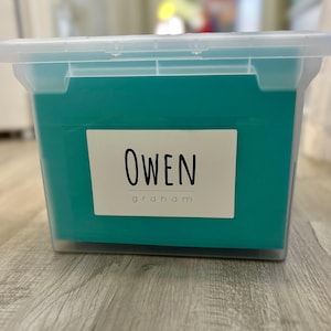 Pode incluir: Uma caixa de armazenamento de plástico transparente com uma tampa turquesa. Um rótulo branco na frente da caixa diz "OWEN graham".