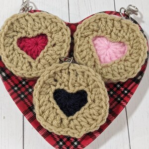 May include: Three crocheted heart-shaped keychains with different colored hearts. The keychains are beige with red, pink, and blue hearts. The keychains are displayed on a red and black plaid heart-shaped plate.