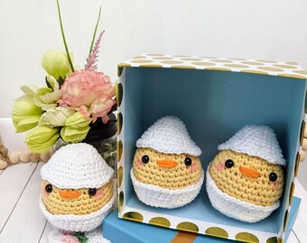 May include: Two crocheted yellow chicks with white hats in a blue and gold polka dot gift box. The chicks are sitting on a blue and gold polka dot box. The box is sitting on a white surface.