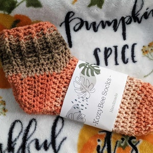 May include: Crocheted socks in orange, brown, and beige colors with a tag that reads "KozyBee Socks - Handmade".