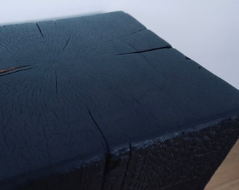 May include: A square, black wooden block with a textured surface and visible wood grain. The top of the block has a series of cracks radiating from the center. The block is a dark charcoal color.