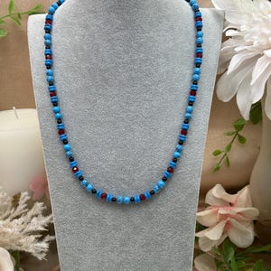 May include: A turquoise, black, and red beaded necklace on a gray mannequin. The necklace is made of small, round beads.