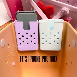 Op de afbeelding: Twee telefoonhouders, &eacute;&eacute;n lavendelkleurig en &eacute;&eacute;n wit, met respectievelijk zwarte en turquoise stippenpatronen. De lavendelhouder bevat een grijze telefoonhoes. De tekst "FITS IPHONE PRO MAX" wordt in het zwart weergegeven. De houders zitten in een oranje container.