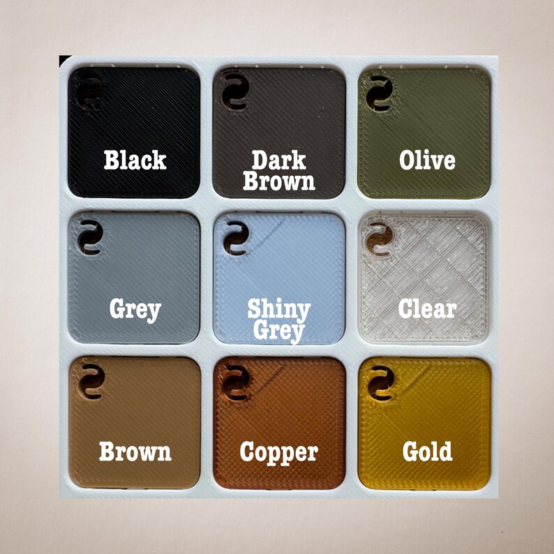 Op de afbeelding: Een raster van negen vierkante kleurstalen, elk met een kleurnaam. Kleuren zijn onder meer zwart, donkerbruin, olijfgroen, grijs, glanzend grijs, helder, bruin, koper en goud. Elke staal heeft een kleine, gebogen uitsparing.