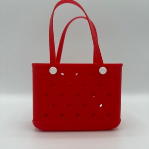 Puede incluir: Bolsa de mano de silicona roja con asas y botones blancos. La bolsa tiene un patr&oacute;n de peque&ntilde;os agujeros y la palabra "bogg" est&aacute; impresa en la parte inferior.