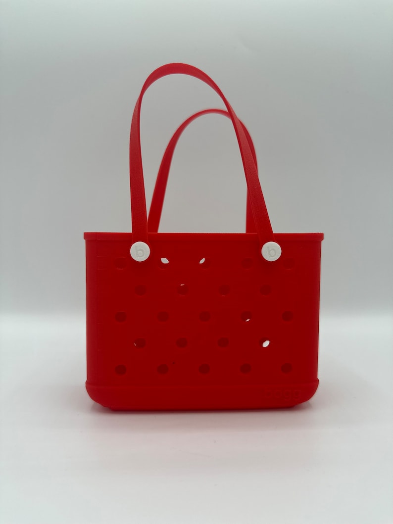 Puede incluir: Bolsa de mano de silicona roja con asas y botones blancos. La bolsa tiene un patr&oacute;n de peque&ntilde;os agujeros y la palabra "bogg" est&aacute; impresa en la parte inferior.