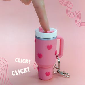 Pode incluir: Caneca de viagem rosa com tampa branca e detalhes prateados, adornada com enfeites em forma de coração rosa. A caneca tem uma alça e um porta-chaves. As palavras "CLICK! CLICK!" estão impressas na lateral.