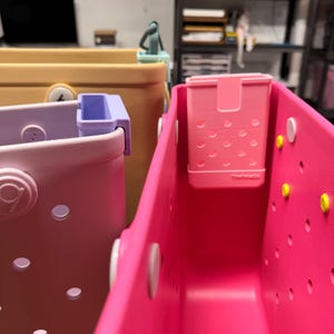 Op de afbeelding: Close-up van verschillende kleurrijke draagtassen in roze, lavendel en geel. Elke tas heeft een bijpassende clip en een kleine, geperforeerde roze container aan de binnenkant. De tassen hebben een cirkelvormig logo en zijn gemaakt van duurzaam materiaal.