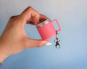 May include: A pink miniature mug keychain with a silver rim and handle. The word "YETI" is printed on the side of the mug. The keychain is attached to a silver clasp.