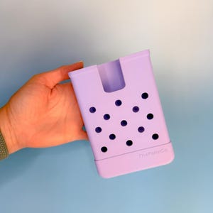 Op de afbeelding: Een lavendelkleurige, rechthoekige organizer met een handgreepachtige uitsparing en een reeks cirkelvormige gaten. De onderkant van de organizer heeft een iets donkerdere lavendelbasis. De tekst "TheFellaCo" staat onderaan.