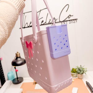 Op de afbeelding: Een lichtpaarse draagtas met een roze strik en een afneembare lavendelkleurige telefoonhouder. De tas heeft een stippenpatroon en een stevige handgreep. De tas wordt vastgehouden door een persoon die een beige trui draagt.