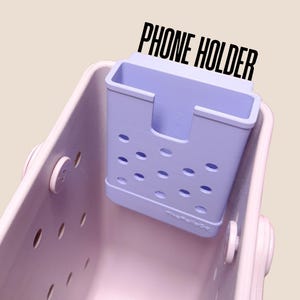Op de afbeelding: Een lichtpaarse telefoonhouder met een rechthoekige vorm en ovale uitsparingen. De tekst "PHONE HOLDER" is in het zwart boven de houder gedrukt. De houder zit in een lichtpaarse draagtas.