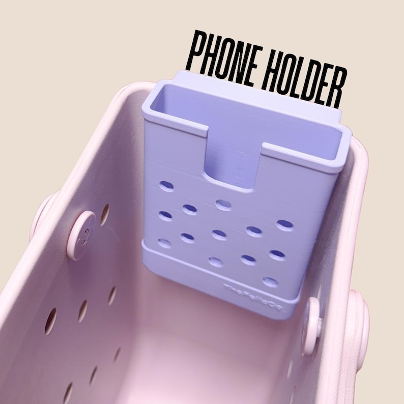 Op de afbeelding: Een lichtpaarse telefoonhouder met een rechthoekige vorm en ovale uitsparingen. De tekst "PHONE HOLDER" is in het zwart boven de houder gedrukt. De houder zit in een lichtpaarse draagtas.