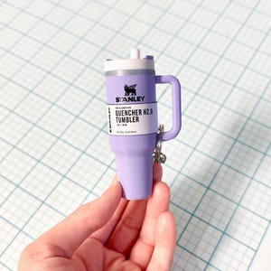 Może przedstawiać: Miniaturowy lawendowy kubek Stanley Quencher H2.0 z białą pokrywką i uchwytem. Kubek ma logo Stanley i informacje o produkcie z przodu. Srebrny brelok do kluczy jest przymocowany do uchwytu.