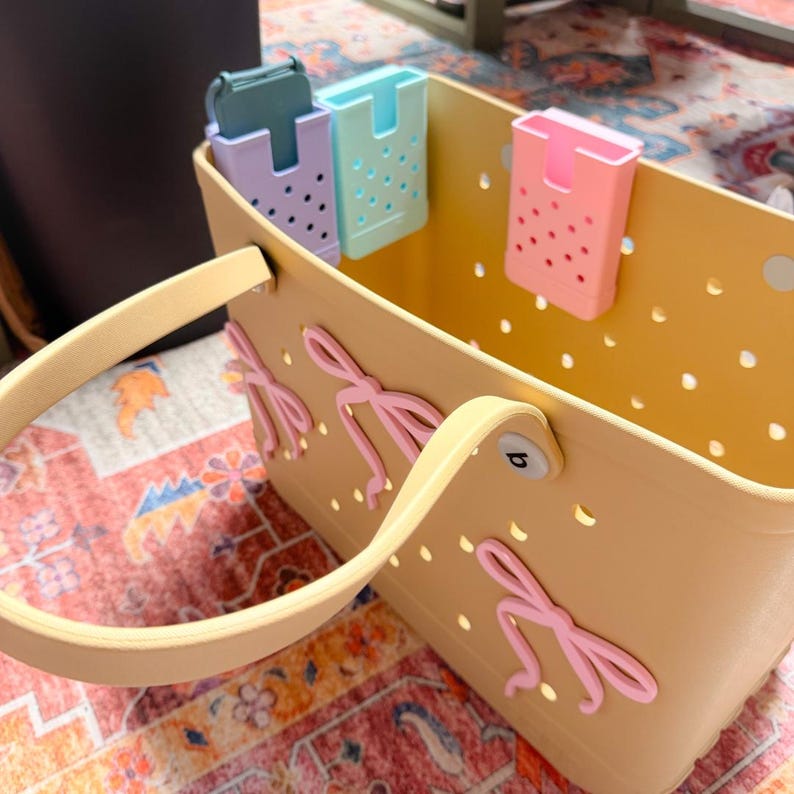 Op de afbeelding: Een lichtgele draagtas met een bijpassende handgreep en roze strikaccenten. De tas heeft een reeks kleine gaatjes en bevat drie pastelkleurige clip-on organizers in groen, blauw en roze. De tas staat op een patroonkleed.