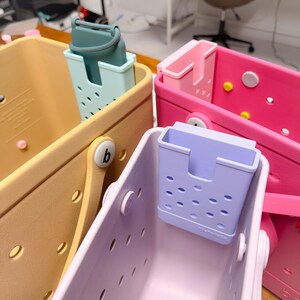Op de afbeelding: Een verzameling kleurrijke, geperforeerde draagtassen en bijpassende bakjes. De tassen zijn in tinten geel, roze en lavendel, met bijpassende bakjes in blauw en paars. Elke tas heeft een ronde knoopsluiting.