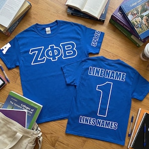 May include: Two royal blue t-shirts with white lettering. One shirt displays "ZΦB" and "A" on the sleeve. The other shirt has "LINE NAME", the number "1", and "LINES NAMES" printed on the back. Books and a coffee cup are in the background.
