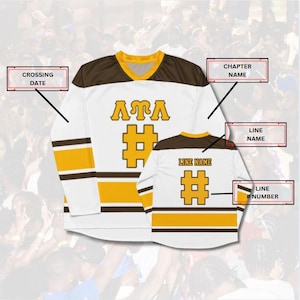 Op de afbeelding: Wit lange mouwen shirt met bruine en gouden accenten. Het toont de letters "ATA" en een gouden hashtag-symbool. Het shirt heeft een V-hals en strepen op de mouwen. De achterkant van het shirt heeft ook een hashtag-symbool en de tekst "LINE NAME".