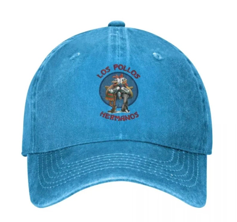 May include: A blue denim baseball cap with a distressed look. The front of the cap features a circular embroidered patch with the text "Los Pollos Hermanos" and a cartoon image of two chickens.