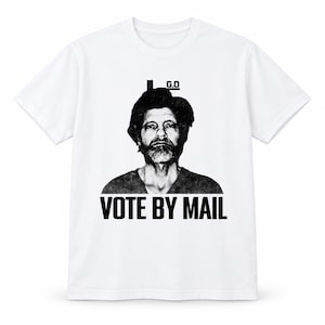 Op de afbeelding: Een wit T-shirt met korte mouwen voorzien van een zwart-witte grafische print van een persoon met een baard, met de tekst G.O boven het hoofd en de woorden VOTE BY MAIL in vette zwarte hoofdletters onder het portret gedrukt.