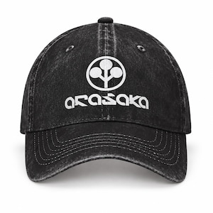 Puede incluir: Una gorra de béisbol negra con un acabado desgastado y lavado. La parte frontal presenta un logotipo circular blanco con un icono de árbol de tres círculos sobre el texto blanco Arasaka en una fuente angular única. Tiene visera curva.