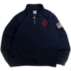Könnte beinhalten: Marineblaues Sweatshirt mit Reißverschluss am Kragen. Es hat ein rotes, gesticktes Logo und einen kleinen Aufnäher mit der amerikanischen Flagge auf dem Ärmel. Das Sweatshirt hat eine lockere Passform und einen gerippten Kragen und Bündchen.