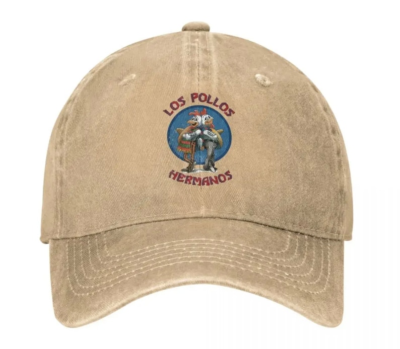 May include: A tan baseball cap with a distressed look. The front of the cap features a circular patch with the text "Los Pollos Hermanos" in red and blue lettering. Inside the circle is a cartoon image of two chickens.