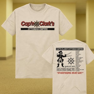 Pode incluir: Uma t-shirt bege de manga curta com o texto vermelho e preto Cap'n Clark's OTTOMAN EMPIRE na frente. As costas exibem um gráfico de pirata, um leme, uma lista de móveis, o número 408-357-2875 e o texto EVERYTHING MUST GO!!