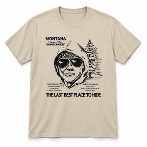 Op de afbeelding: Een beige t-shirt met korte mouwen en een zwarte grafische print van een persoon in een hoodie met zonnebril. De tekst luidt MONTANA Home of the UNABOMBER, MONTANA'S STATE SLOGANS en THE LAST BEST PLACE TO HIDE met een dennenboom.