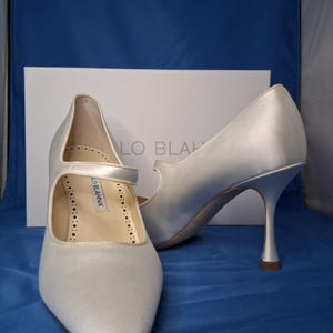 Puede incluir: Un par de zapatos de tacón alto de satén marfil con punta y correa Mary Jane. Los zapatos tienen tacones delgados y se exhiben frente a una caja blanca con el nombre de la marca. El fondo es azul liso.