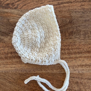 May include: A cream-coloured, crocheted baby bonnet with a rounded crown and a chin tie. The bonnet is made of a textured yarn and is shown on a wooden surface. The tie is made of the same yarn as the bonnet.