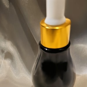 May include: A black glass bottle with a gold-colored collar and a white dropper. The bottle has a rounded shape and a glossy finish. The dropper is white and has a rounded tip. The bottle is likely for essential oils or serums.