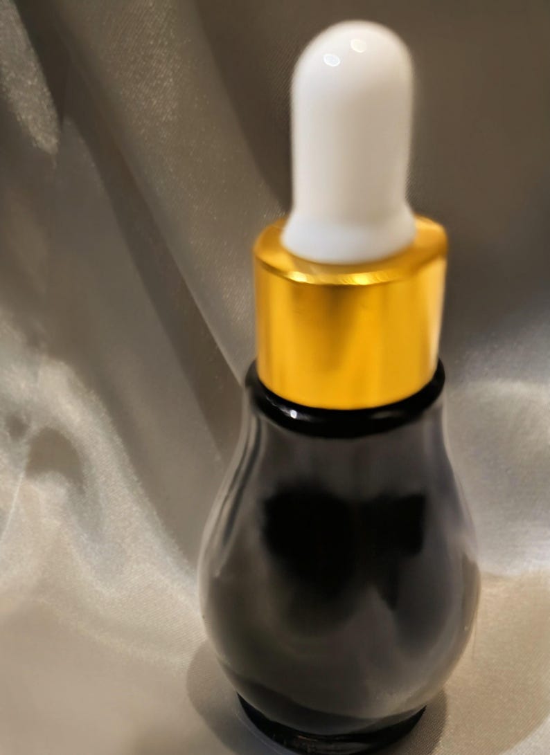May include: A black glass bottle with a gold-colored collar and a white dropper. The bottle has a rounded shape and a glossy finish. The dropper is white and has a rounded tip. The bottle is likely for essential oils or serums.