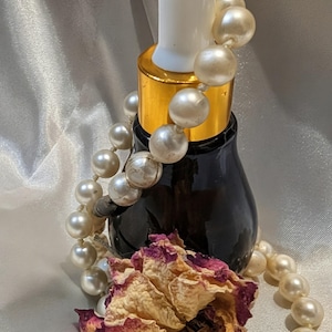 May include: A dark glass bottle with a white dropper and gold collar, adorned with a pearl necklace and a dried rose. The bottle is likely for essential oils or serums, creating a vintage aesthetic.
