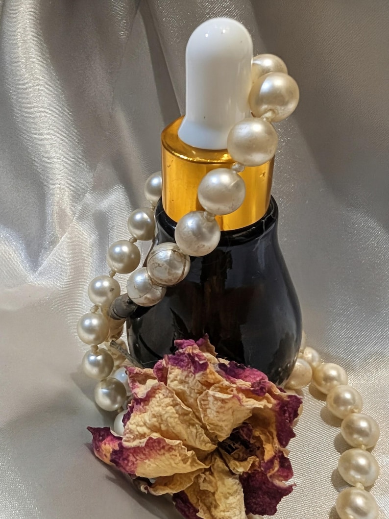 May include: A dark glass bottle with a white dropper and gold collar, adorned with a pearl necklace and a dried rose. The bottle is likely for essential oils or serums, creating a vintage aesthetic.