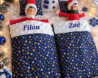 Puede incluir: Dos muñecos de elfo acostados en sacos de dormir azul marino a juego con patrones de estrellas doradas y blancas. El borde de felpa blanca de los sacos lleva bordados los nombres Filou y Zoé. Un muñeco de nieve de peluche con gorro y bufanda azules se sitúa entre ambos, rodeado de adornos navideños.