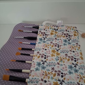 Peut inclure: Un étui à pinceaux en tissu avec un motif floral dans des tons bleu, jaune et rose sur fond blanc, et un intérieur à pois lavande. L'étui contient neuf pinceaux assortis dotés de manches noirs et de poils blancs.