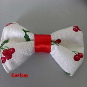 Peut inclure: Un nœud papillon blanc orné de motifs de cerises rouges et de feuilles vertes, avec un ruban rouge vif au centre. Le mot "Cerises" est écrit en rouge en bas de l'image.