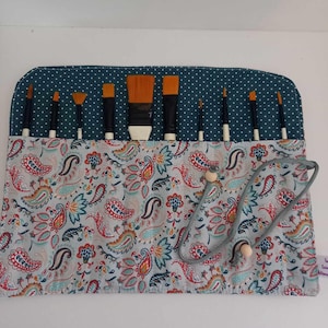 Peut inclure: Un étui enroulable en tissu bleu sarcelle et blanc avec un motif paisley et un cordon de serrage gris. L'étui est rempli d'un ensemble de pinceaux à peinture avec des poils orange et blancs.