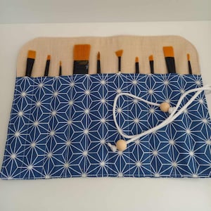 Peut inclure: Un étui enroulable en tissu bleu et blanc à motif avec un cordon de serrage blanc et des perles en bois. L'étui est rempli d'un ensemble de pinceaux.